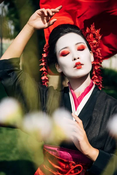 Selective focus of tree branches and beautiful gesturing geisha with red cloth on background in sunlight — Stock Photo