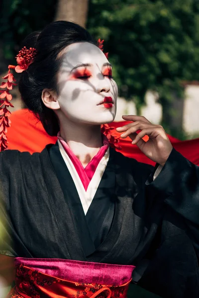 Fuoco selettivo di rami d'albero e bella geisha con gli occhi chiusi e panno rosso sullo sfondo alla luce del sole — Foto stock