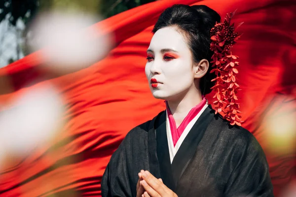 Selective focus of tree branches and beautiful geisha looking away with red cloth on background in sunlight — Stock Photo