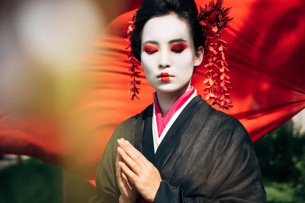 Enfoque selectivo de ramas de árboles y hermosas geishas con gesto de saludo y tela roja en el fondo a la luz del sol - foto de stock