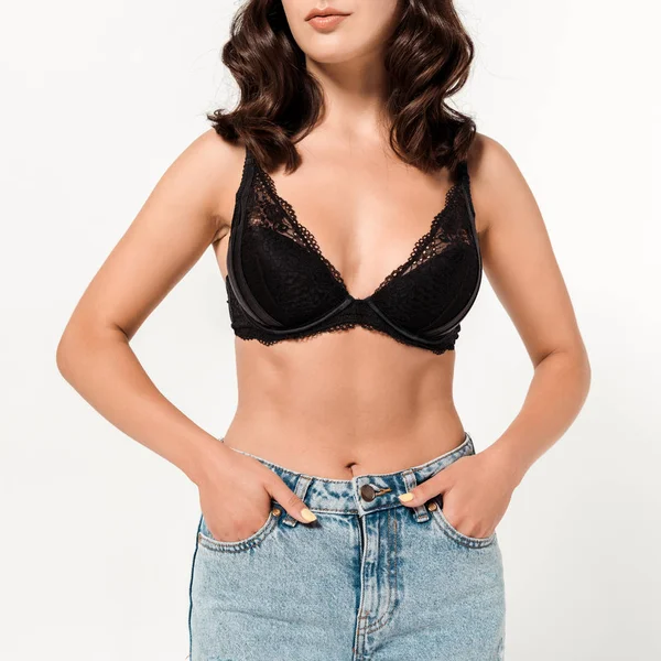 Cropped view of girl in lace bra and blue jeans standing with hands in pockets on white — Stock Photo