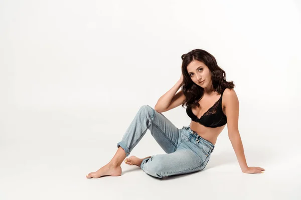 Attractive woman with barefoot sitting in blue jeans and touching hair on white — Stock Photo