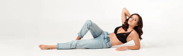 Panoramic shot of attractive woman with barefoot lying in blue jeans and touching hair on white — Stock Photo