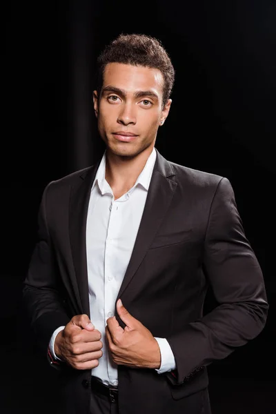 Handsome mixed race man looking at camera while touching suit on black — Stock Photo