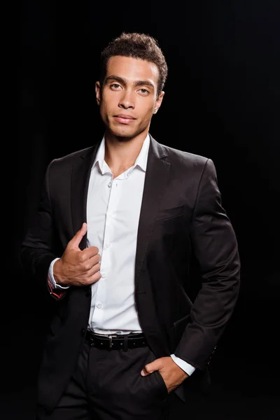 Handsome mixed race man touching suit while standing with hand in pocket isolated on black — Stock Photo