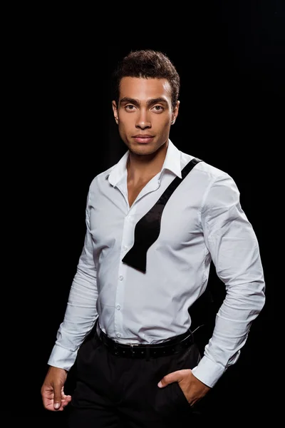 Handsome mixed race man looking at camera while standing with hand in pocket isolated on black — Stock Photo