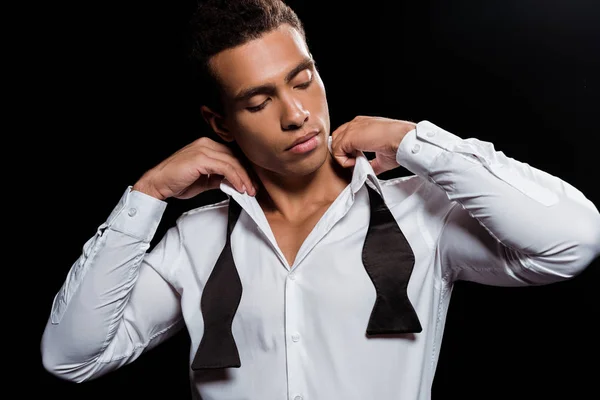 Handsome mixed race man standing and touching tie isolated on black — Stock Photo
