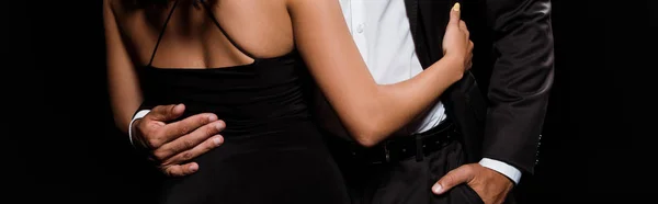Tiro panorâmico de mulher jovem abraçando homem de pé com a mão no bolso isolado no preto — Fotografia de Stock