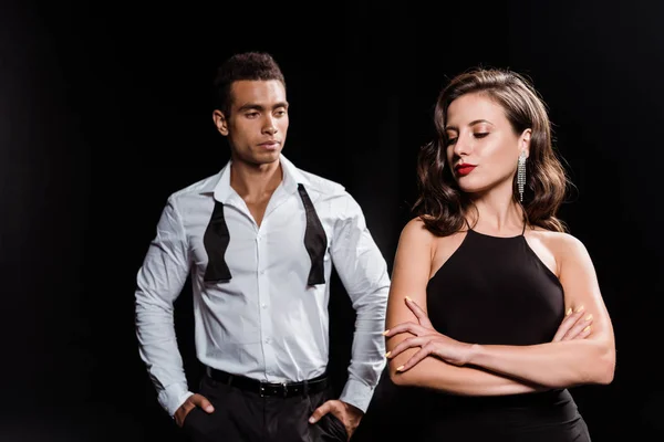 Handsome mixed race man standing with hands in pockets near woman with crossed arms isolated on black — Stock Photo