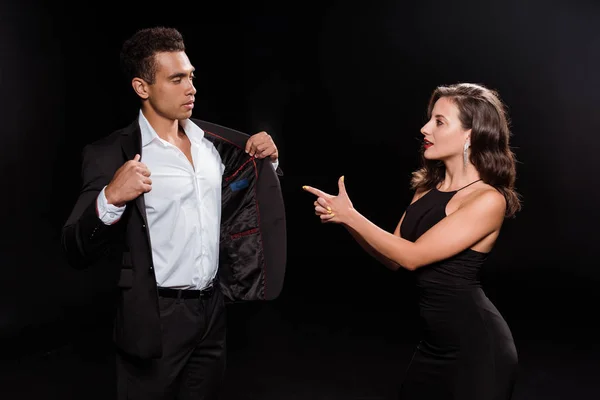 Приваблива дівчина вказує пальцем на змішану расу чоловік торкається костюма ізольовано на чорному — стокове фото