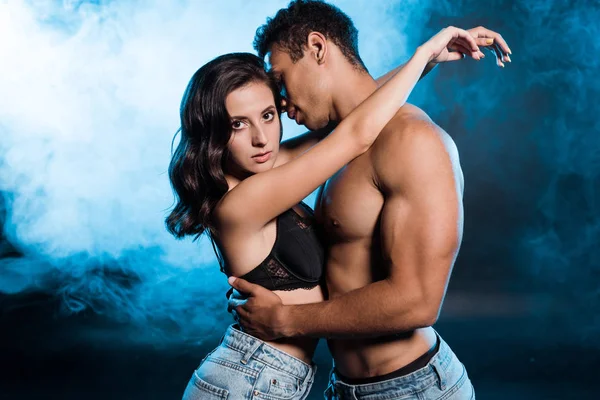 Mestizo hombre abrazando y mirando sexy chica en sujetador y jeans en azul con humo - foto de stock