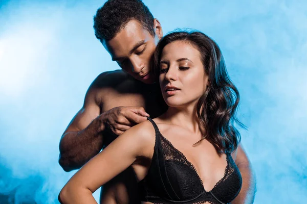 Schöner bi-rassischer Mann, der einen schwarzen Spitzenbüstenhalter von sexy Mädchen auf blauem Grund mit Rauch berührt — Stockfoto