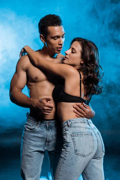 Sexy mixta raza hombre abrazando y mirando a chica en sujetador y jeans en azul con humo - foto de stock