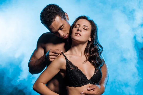 Shirtless mixed race man touching bra on sexy woman on blue with smoke — Stock Photo