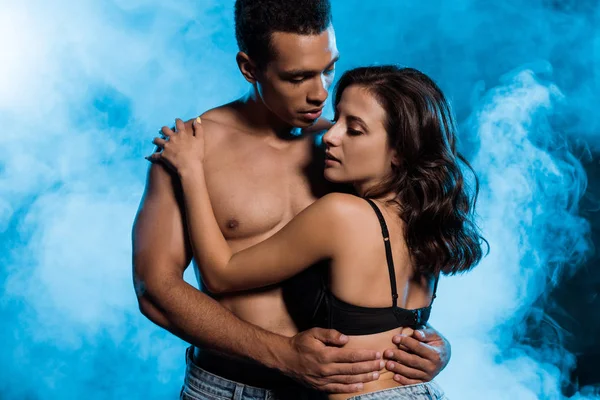 Sin camisa mezclado raza hombre abrazando sexy mujer en sujetador en azul con humo — Stock Photo