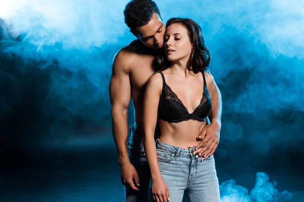 Handsome mixed race man hugging attractive girl in lace black bra on black with blue smoke — Stock Photo