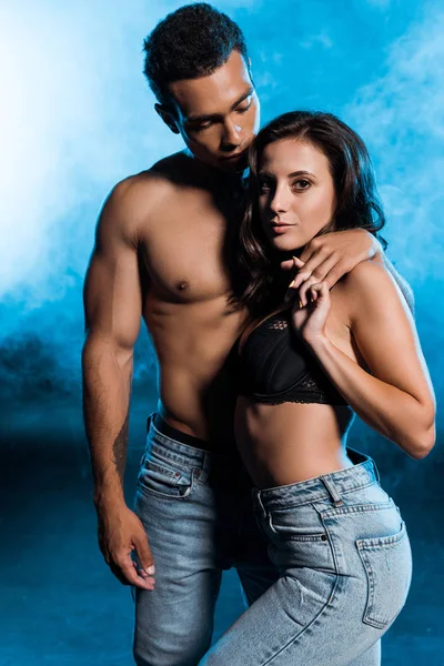 Shirtless mixed race man holding hands with sexy girl on blue with smoke — Stock Photo