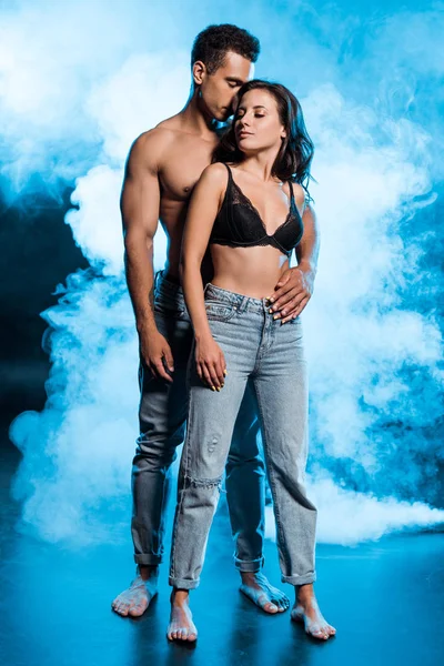 Sem camisa misto raça homem abraçando sexy menina em jeans jeans de pé no azul com fumaça — Fotografia de Stock