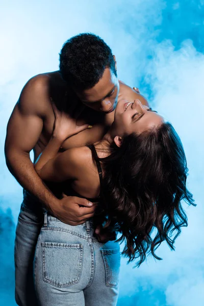 Shirtless mixed race man hugging happy girl in denim jeans standing on blue with smoke — Stock Photo