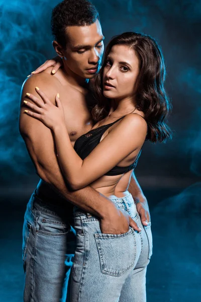 Muscular mixed race man hugging girl in jeans and putting hands in pockets on blue with smoke — Stock Photo