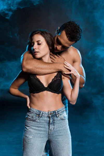 Muscular bi-racial man hugging girl in jeans and bra standing with hand on hip on blue with smoke — Stock Photo