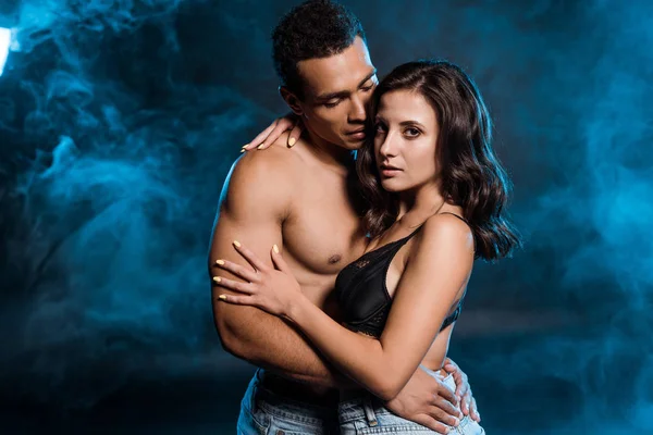 Muscular bi-racial man hugging young woman in jeans and bra on black with smoke — Stock Photo