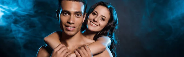 Panoramic shot of happy woman hugging muscular mixed race man on black with blue smoke — Stock Photo