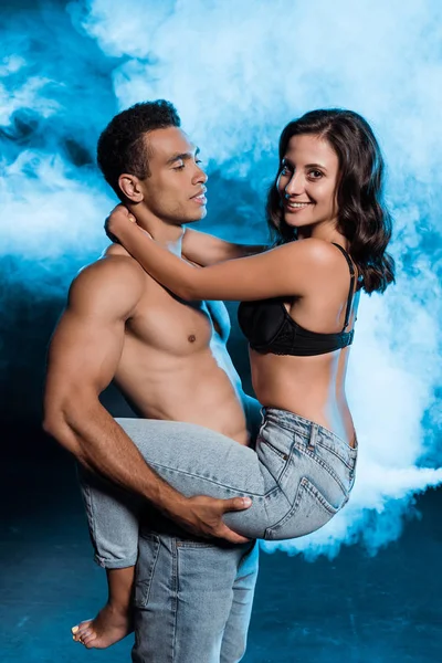 Muscular mixed race man holding in arms happy girl in denim jeans on blue with smoke — Stock Photo