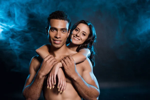 Giovane donna allegra abbracciando muscolare uomo razza mista su nero con fumo blu — Foto stock