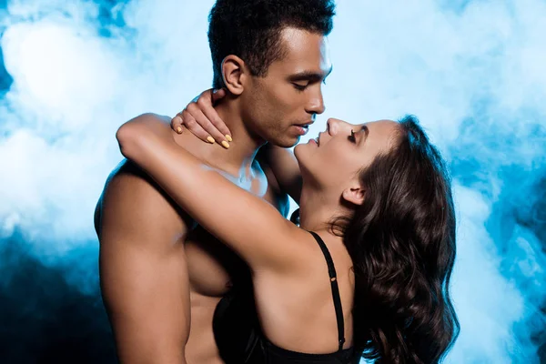 Hemdlos Mixed Race Mann umarmt junge attraktive Frau in Spitzen-BH auf blau mit Rauch — Stockfoto