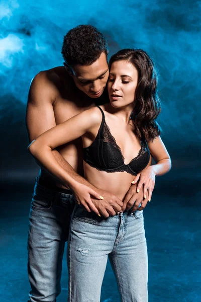 Sexy shirtless man hugging young woman standing in jeans and lace bra on blue with smoke — Stock Photo