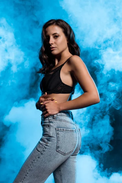 Young woman in lace bra and denim jeans on blue with smoke — Stock Photo