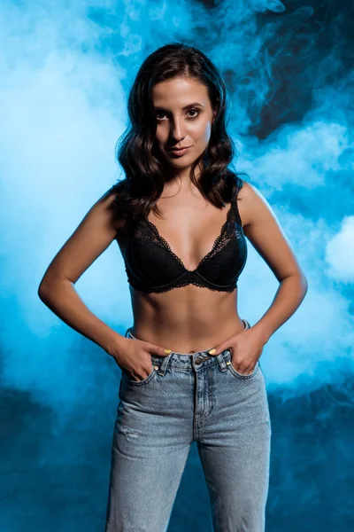 Sexy giovane donna in pizzo reggiseno in piedi con le mani in tasca su blu con il fumo — Foto stock