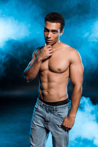 Sexy mixed race man standing in denim jeans and touching face on blue with smoke — Stock Photo