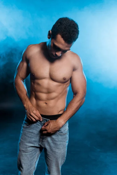 Muscular mixed race man touching denim jeans on blue with smoke — Stock Photo