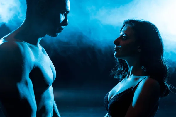 Side view of young woman in bra looking at sexy muscular mixed race man on blue with smoke — Stock Photo