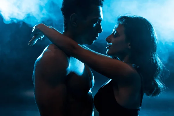 Side view of young attractive woman in bra hugging sexy muscular mixed race man on blue with smoke — Stock Photo