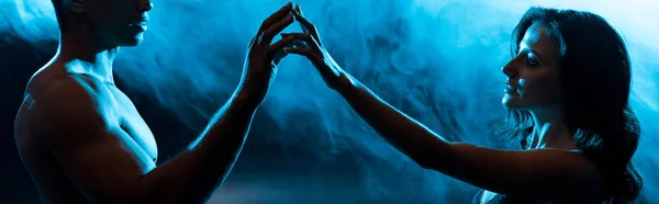 Panoramic shot of young woman in bra touching hands with sexy muscular mixed race man on blue with smoke — Stock Photo