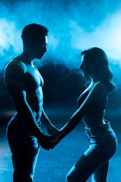 Вид сбоку молодой женщины в лифчике, держащейся за руки с сексуальным мускулистым мужчиной на синем с дымом — стоковое фото