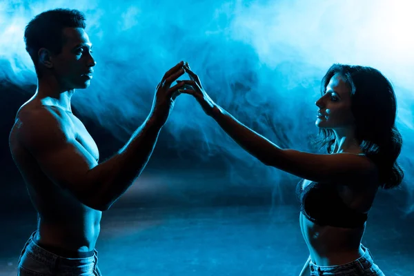 Young woman in bra touching hands with sexy muscular mixed race man on blue with smoke — Stock Photo