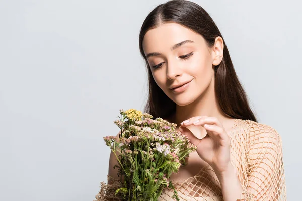 Belle femme souriante en maille beige regardant des fleurs sauvages isolées sur gris — Photo de stock