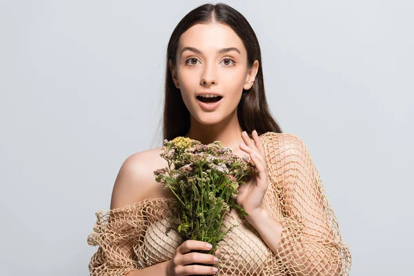 Hermosa mujer conmocionada en malla beige con flores silvestres mirando hacia otro lado aislado en gris - foto de stock