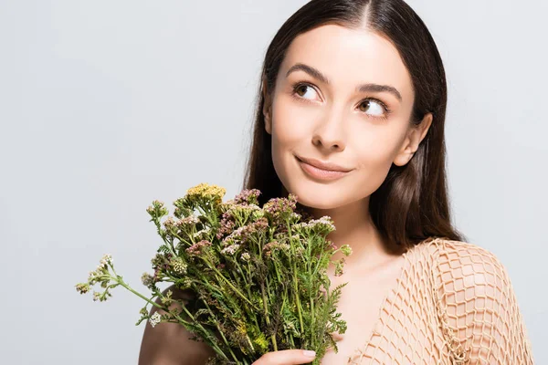 Hermosa mujer sonriente en malla beige con flores silvestres mirando hacia otro lado aislado en gris - foto de stock