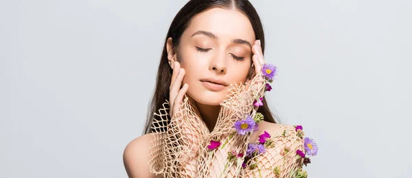 Belle femme aux yeux fermés en maille vêtements beige avec des fleurs violettes touchant visage isolé sur gris — Photo de stock