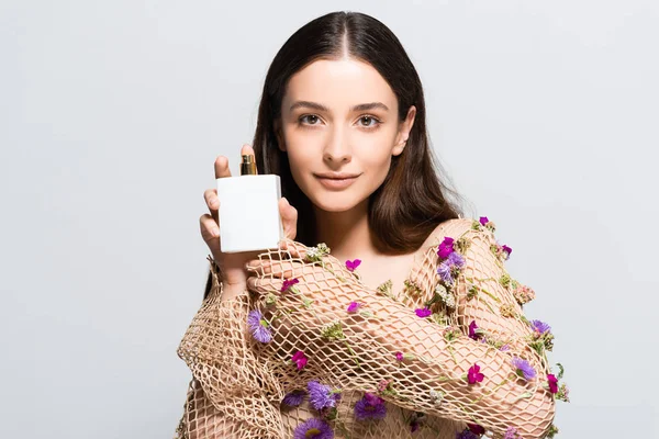 Beautiful woman in mesh clothing with purple flowers holding perfume bottle with copy space and floral scent isolated on grey — Stock Photo