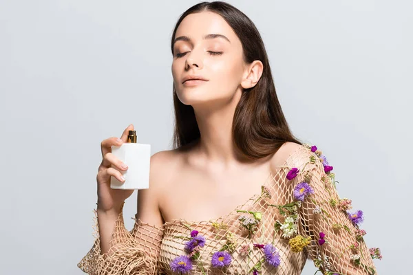 Hermosa mujer en ropa de malla con flores púrpuras rociando perfume con aroma floral en la piel aislada en gris - foto de stock