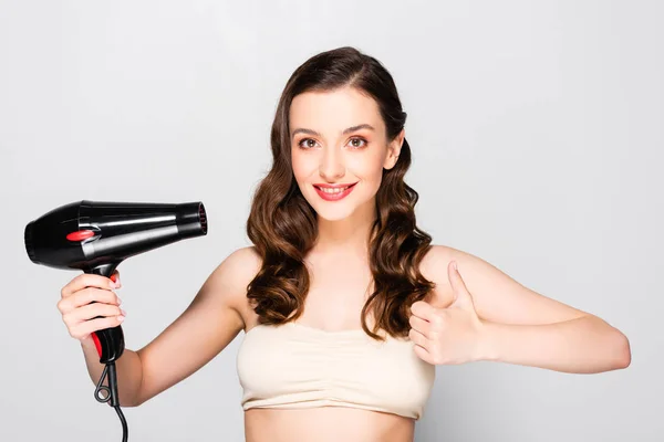 Beautiful brunette woman with curls and makeup holding hairdryer and showing thumb up isolated on grey — Stock Photo