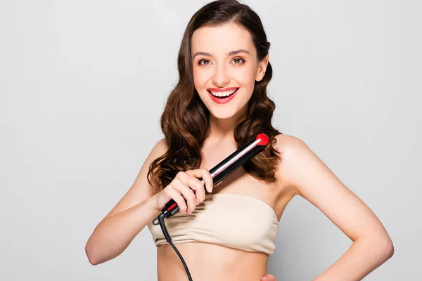 Happy beautiful brunette woman with curls and makeup holding curling iron isolated on grey — Stock Photo