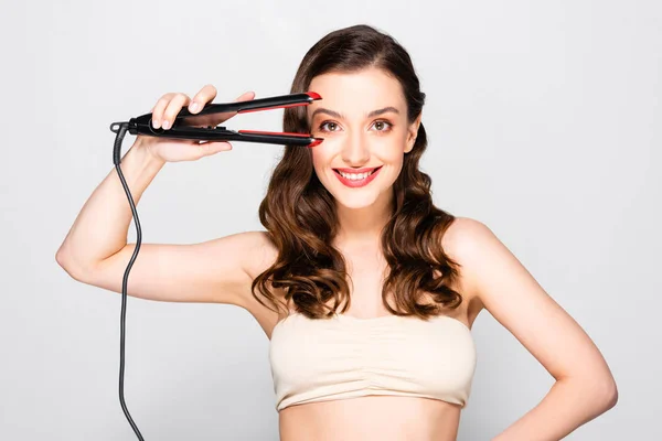 Smiling beautiful brunette woman with curls and makeup holding straightening flat iron isolated on grey — Stock Photo