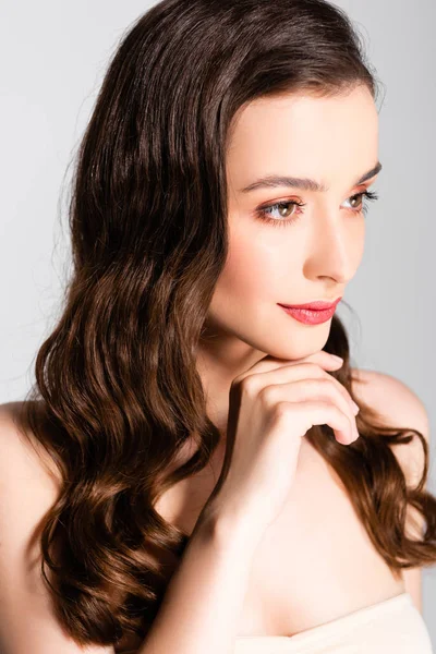 Beautiful brunette woman with curls and makeup looking away with hand near chin isolated on grey — Stock Photo